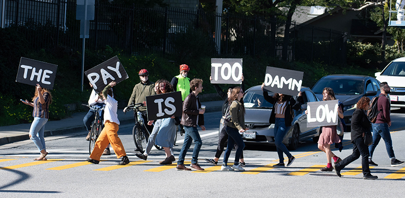 grad students blocking traffic in the street with signs that read "The Pay Is Too Damn Low"