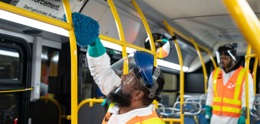 A worker cleaning a yellow pole with disinfectant on a NYC bus with another worker looking on. the workers are wearing orange safety vests, helmets, and transparent visors.
