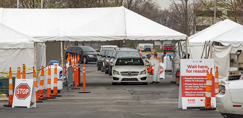 A line of cars coming through a coronavirus testing site in a parking lot. Site has tents.
