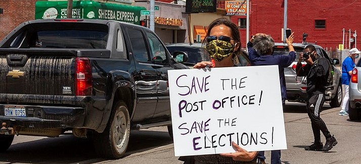 A masked protester holds a sign: "Save the Post Office! Save the Elections!" Behind the person, traffic and a "Coney Island" (diner) across the street