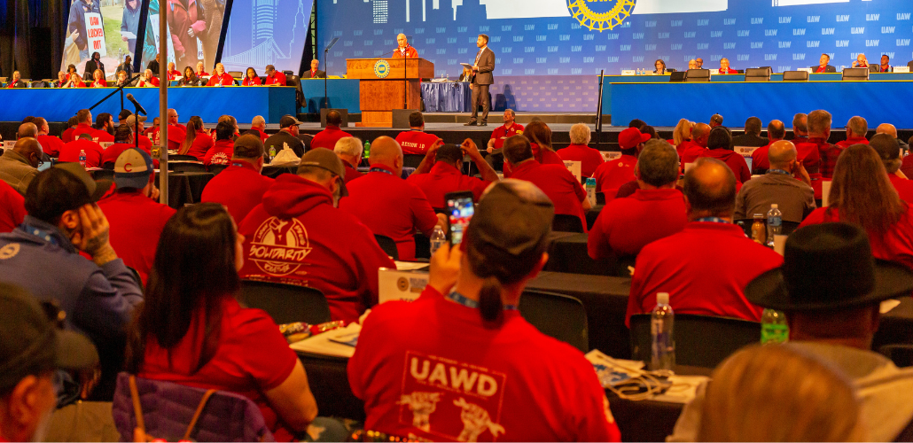 View from back of an auditorium at UAW convention. People in red shirts sit in rows; one red hoodie front and center has UAWD logo on the back. Onstage, Shawn Fain is at the podium, someone in a suit stands near him, and other people mostly in red shirts sit arrayed at long tables on the dais. A big blue screen behind them has UAW logo and intials repeated, and some other images not clearly visible.