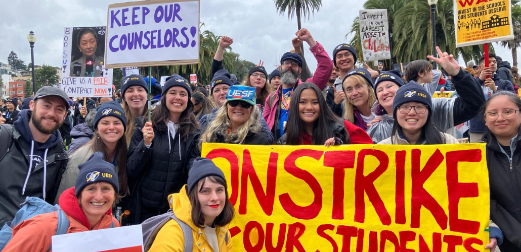 Smiling strikers pose outdoors with a bright yellow banner handpainted "On strike for our schools" in red. Most people visible in this photo appear to be white or Asian women. Many wear UESF hats. Other visible handmade signs say "Boo Su, give us a fair contract now!" "Keep our counselors" and "Invest in oiur largest asset: children." Palm trees are visible against gray sky. Some people raise fists or a victory/peace sign.