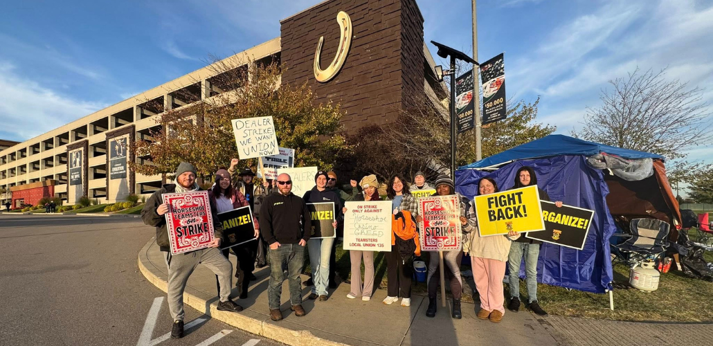A dozen or so workers stand on a corner in from of a parking garage with a giant horseshoe logo on it. They look determined. They carry a mix of hand-lettered and printed signs saying things like "Dealers on strike," "Organize," "Fight back," "Horseshoe Teamsters on strike," and "On strike only against Horseshoe Casino Greed, Teamsters Local 135." Behind them is a canopy that has been insulated with tarps closing some of the sides. Camp chairs and a propane tank are visible. Some wear knitted hats.