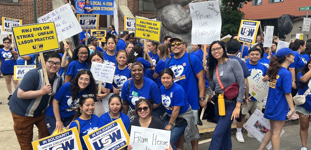 A group of 25 fired-up nurses in royal blue t-shirts poses with signs about striking and safe staffing.