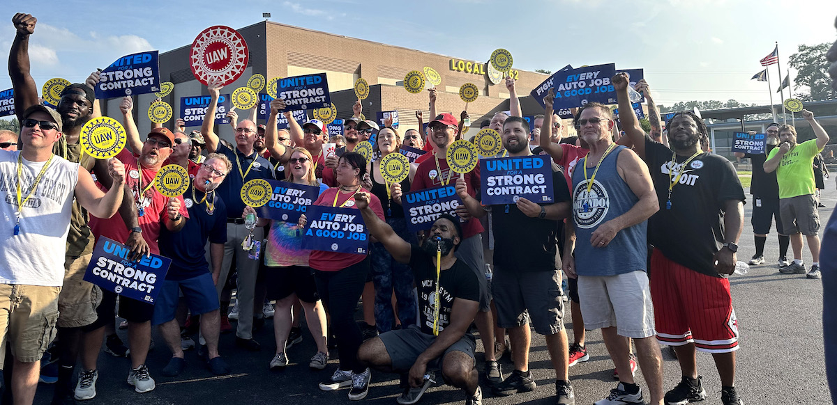 A fired up crowd of about 50 holds round yellow signs saying UAW Local 862 and blue and white signs saying United for a Strong Contract and Every Auto Job a Good Job.