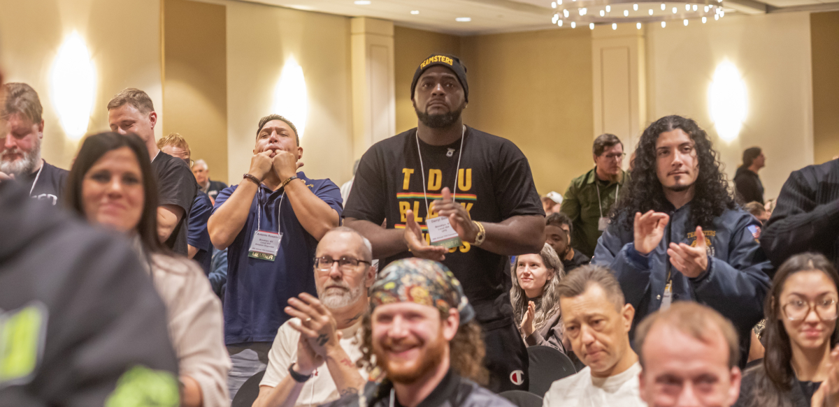 TDU convention attendees are shown sitting and standing, clapping for a speaker, in the hotel ballroom, under a chandelier.