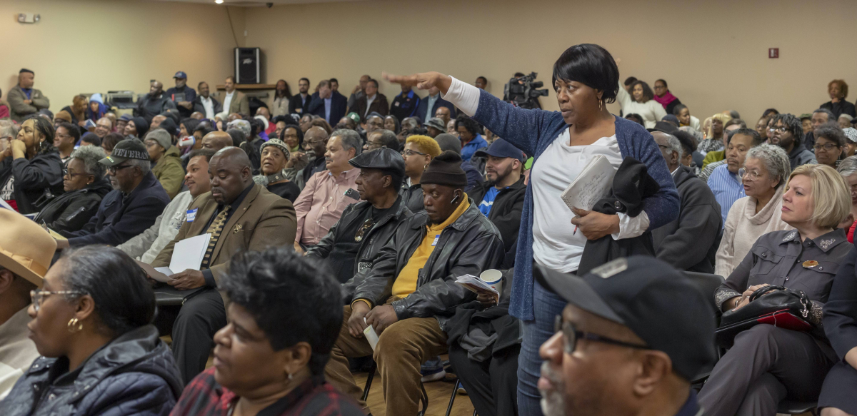 A woman stands up to speak in a big meeting, holding a notebook in one hand.