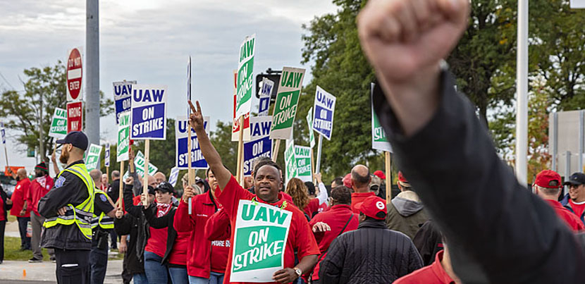 GM workers on a picket line, one work offscreen with arm raised, another centered with sign with arm raised.