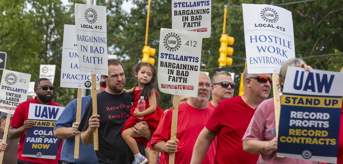 Auto workers march with signs that say “Stellantis Bargining in bad faith” One man is carrying a four-year old child.