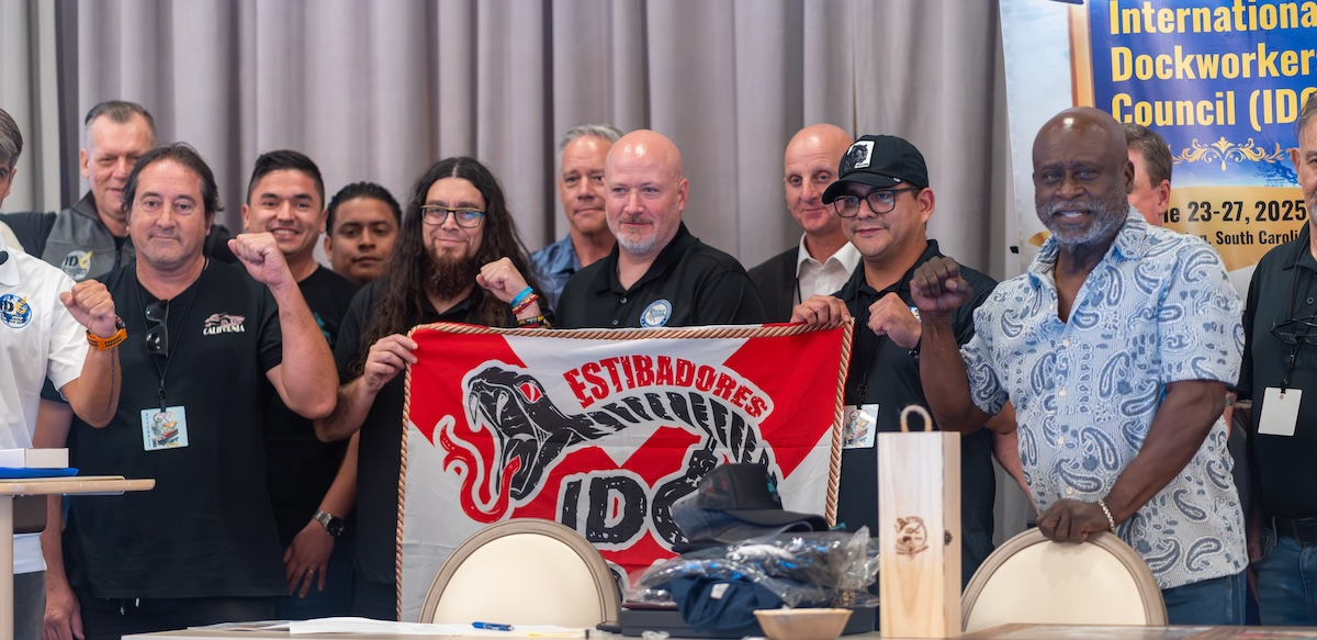 A group of a dozen men of various ages hold a red and white banner that says “Estibadores”