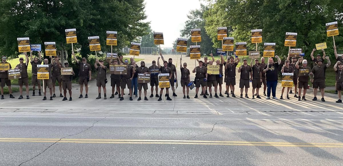 A row of people in UPS outfits with signs seen from far away block the road to some kind of facility, a gate is visible behind them.