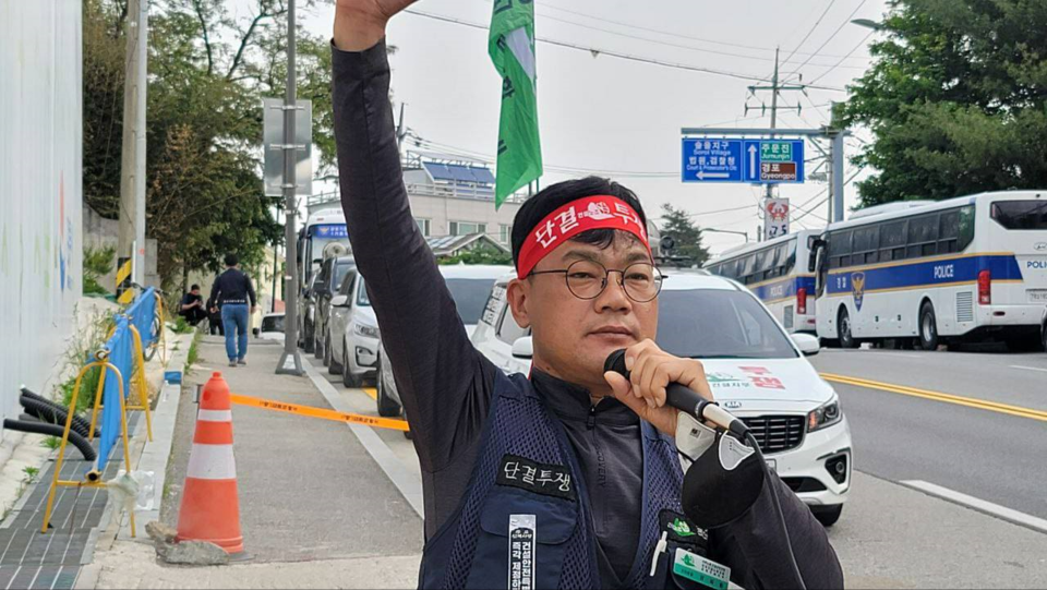 A man, Yang Hoe-dong, walks along a sidewalk wearing a resolute expression. His right arm is in the air, holding a green flag. In his left hand he holds a microphone to his lips. He wears a red headbank with printing on it in Korean, and a vest and badge that also have printed text in Korean. It seems likely he is speaking or leading chants at a union protest.