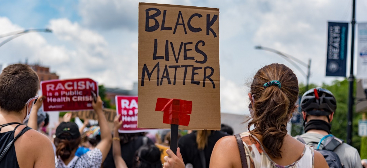 Crowd viewed from back. Sign in foreground, hand-lettered: "Black Lives Matter." Signs in background, printed: "Racism is a public health crisis" with the logo of National Nurses United