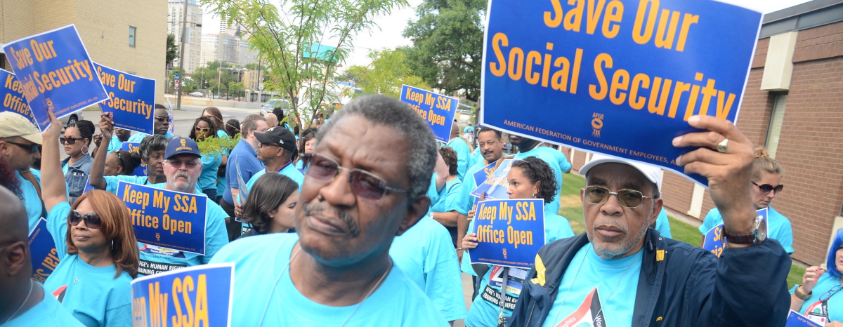 A crowd dressed in light blue T-shirts holds signs saying Save our Social Security and Keep My SSA Office Open.