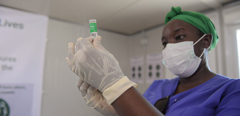 Medical officer in blue scrubs, green bandana, and surgical mask preparing vaccine for administration