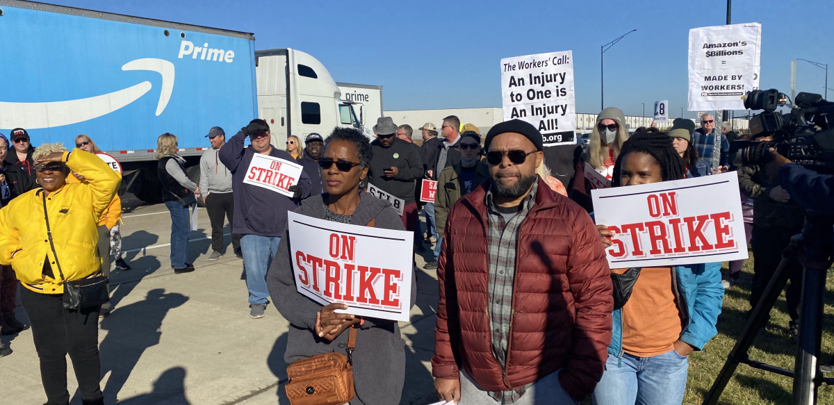 A group of workers and supporters hold signs that read STRIKE in front of a blue Amazon Prime truck.