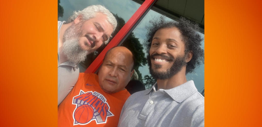 Three workers pose for a group selfie in front of the store. They read as three men of different races. The man in the middle is wearing an orange Knicks T-shirt.