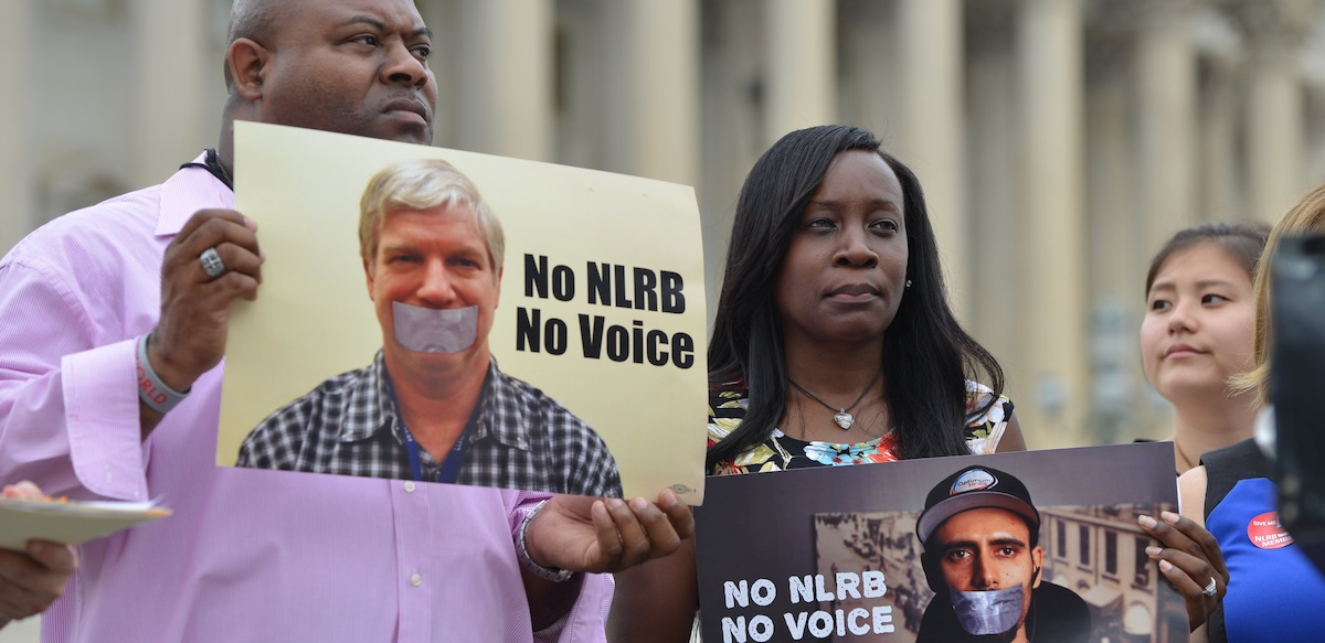 A man and a woman hold signs saying “no NLRB, no voice” with the columns of a government building behind them