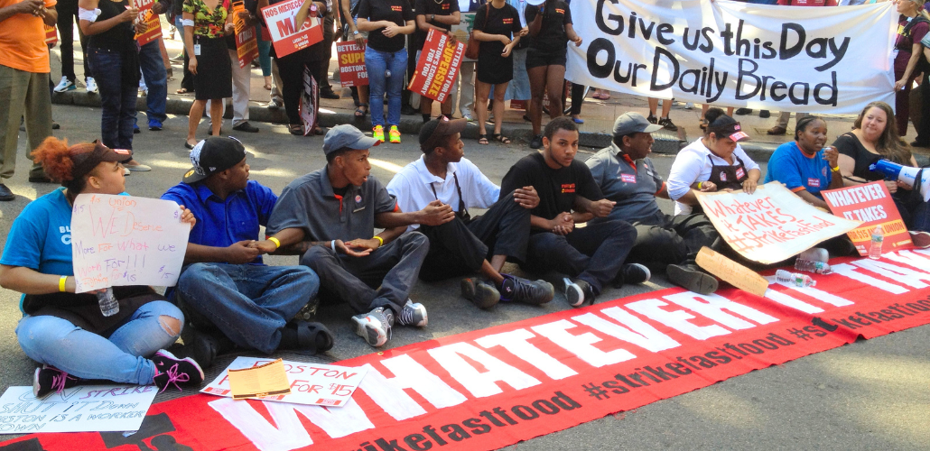 Fast food strikers, mostly young and Black, a mix of genders, sit in an intersection, linking arms. A long red banner laid out in front of them says "Whatever it takes." In the crowd behind them, some people hold a white handlettered banner: "Give us this day our daily bread."