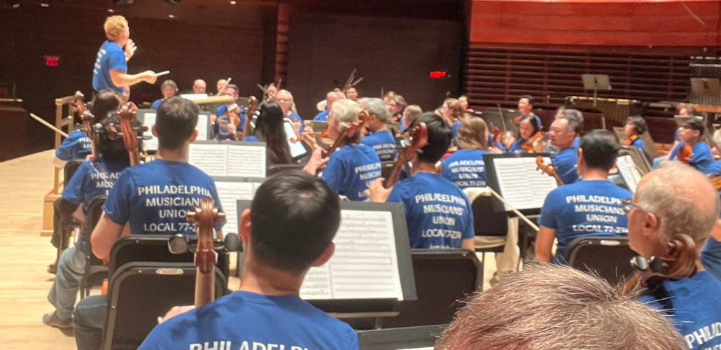 Orchestra musicians on a stage, apparently performing or rehearsing (conductor's baton is raised). Everyone, including the conductor, is wearing matching blue T-shirts that say "Philadelphia Musicians Union Local 77-274" on the back.