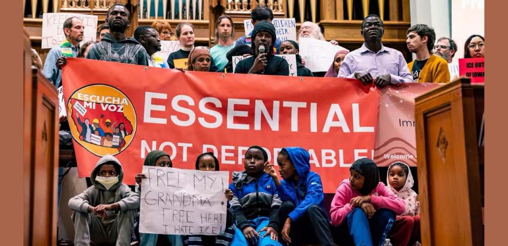 A diverse and serious crowd of people stands close together inside what looks like a church, in front of organ pipes, holding a big printed red banner that says "Essential not deportable" with the logo of Escucha Mi Voz. The person in the middle is speaking into a mic. Several kids sit in front and one holds a hand-lettered sign: "Free my grandma! Free her! I hate ICE!"