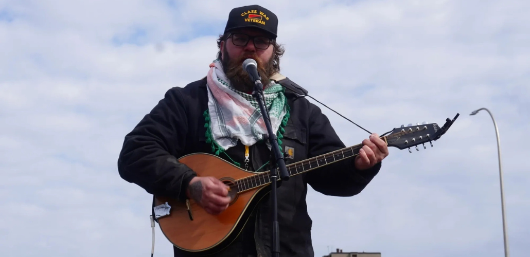 Emmett Doyle, a bearded white man wearing a "Class war veteran" hat, a keffiyeh, and a Carhartt jacket, plays an Irish bouzouki. He is outdoors, silhouetted against a blue sky with thin white clouds.
