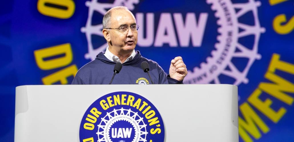 Shawn Fain, a white man with glasses, speaks at a podium. Behind him (and on the podium) is the UAW logo in white on a blue background. Yellow letters around it say (partly obscured) "Our Generation's Defining Moment."