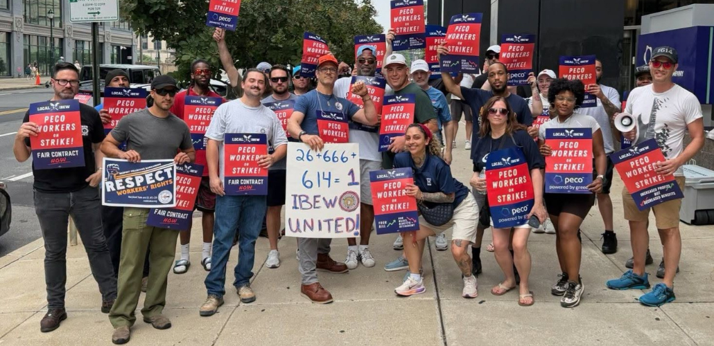 A group of workers pose for the camera on a city sidewalk. They are diverse in race, gender, and age. Many hold printed red and blue signs that say "PECO workers on strike!" One holds a big hand-drawn sign: "26+666+614=1. IBEW united!"
