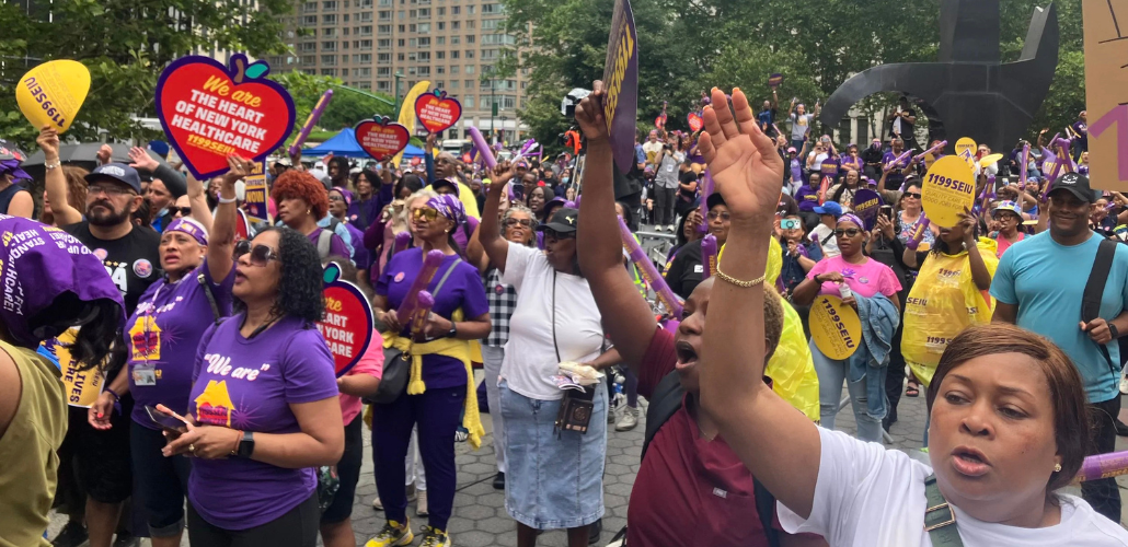 A large crowd of people, mostly Black women, rallies in Manhattan's Foley Square. Many wear purple 1199 T-shirts. Some hold yellow 1199 signs, or red ones shaped like hearts with the message "We are the heart of New York healthcare." The mood is active, with many arms reaching up into the air and mouths opening to speak, chant, or sing.