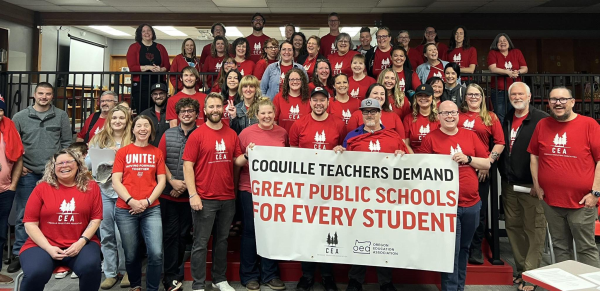 A group of dozens of teachers wearing red shirts gather behind a union banner.