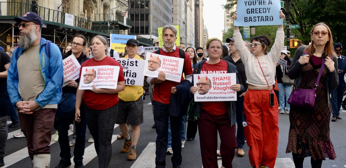 Eight people walk towards the camera, three with signs that say, “shame, Russel Vought”