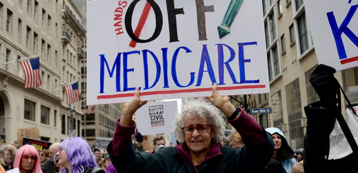 A woman in a big crowd holds up a sign saying ‘hands off Medicare’