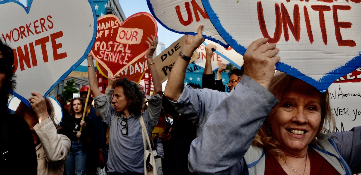 Protesters crowd towards the camera with heart-shaped signs saying “workers unite” and “hands off our postal service”