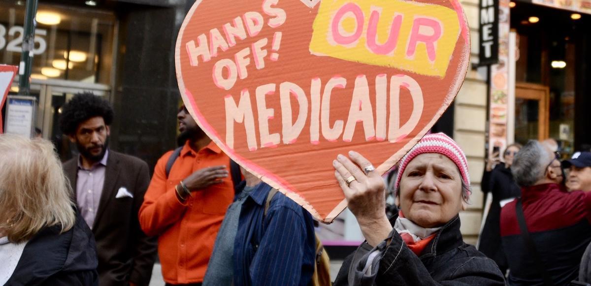 An older person holds up a handmade orange sign saying “Hands off our Medicaid”