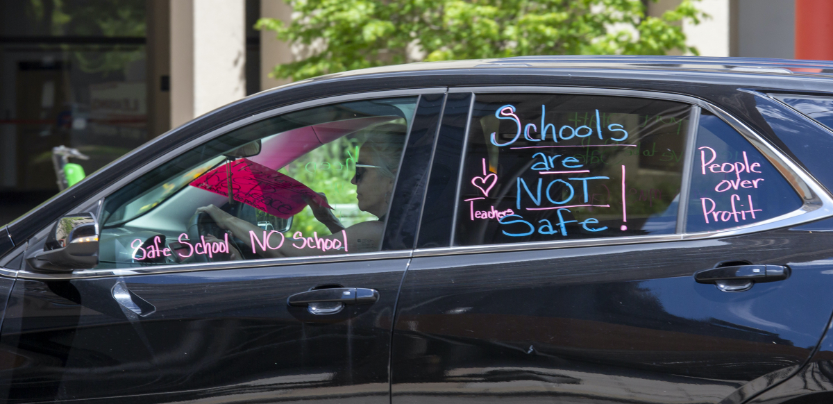 A woman drives a car with the words "Schools are not safe!" "I love teachers" and "People over profit" painted on the windows