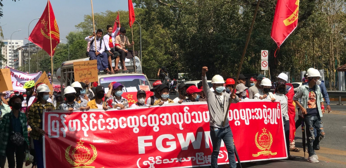 Dozens of workers march behind a red banner in Myanmar. Some carry red flags.
