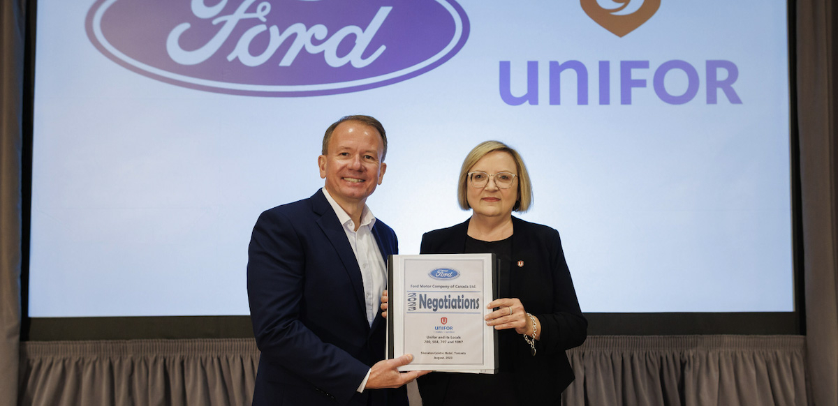 A man and a woman in suits stand holding a binder together in front of a screen that has Ford and Unifor logos projected on it.