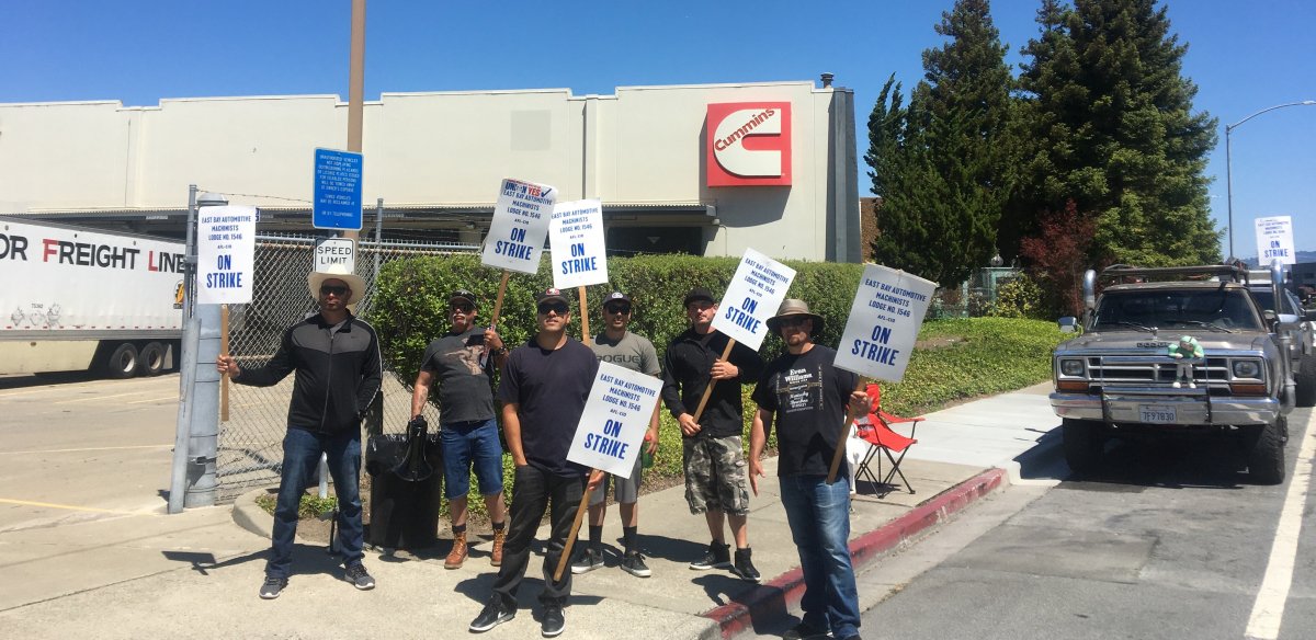 Members of Machinists Local 1546 stand in front of their workplace holding picket signs.