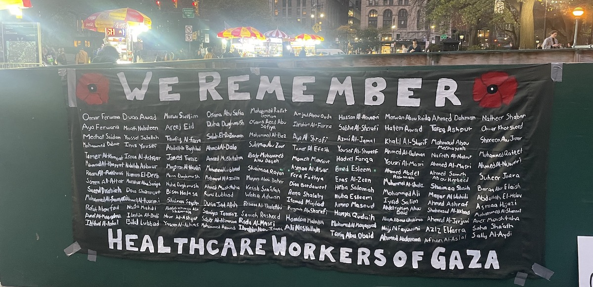 A black banner with white letters lists 160 health care workers killed in Gaza. It is titled “We Remember”.