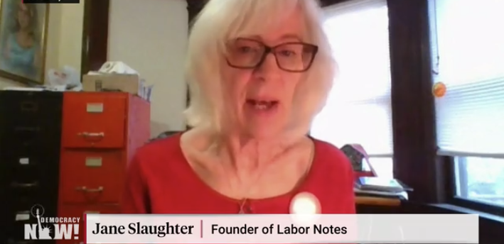 A blond woman in a red scoopneck shirt and black framed glasses speaks to the camera.