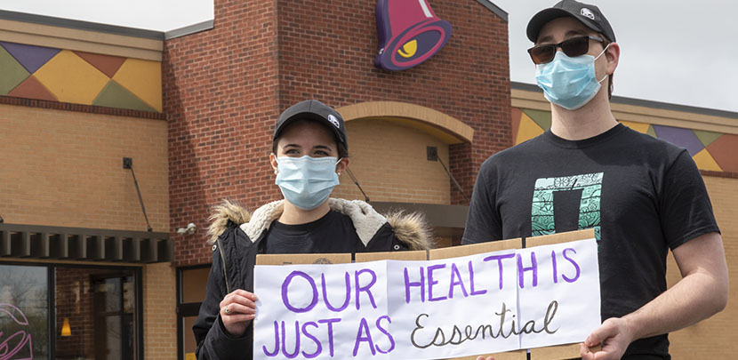 Two taco bell workers with masks holding up a sign in front of a taco bell that says "our health is just as essential."