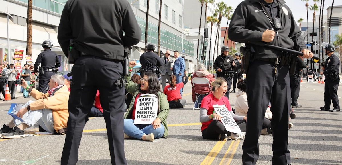 Several workers sit in the street with signs saying “Kaiser Don’t Deny Mental Health”--they are surrounded by police.