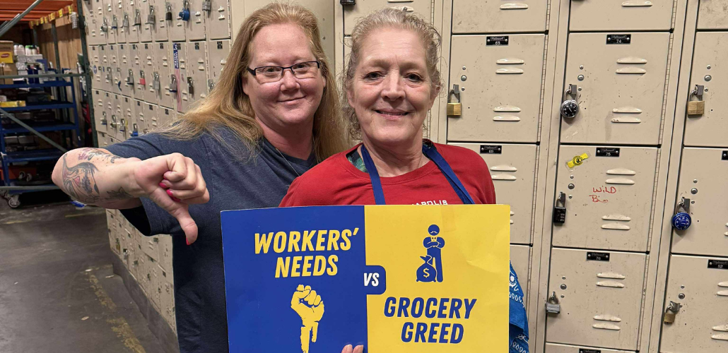 Two white women stand together in front of a wall of lockers, presumably the non-public area of a grocery store, holding a printed blue and yellow sign that reads "Workers' needs vs. grocery greed," with symbols of a fist and Mr. Moneybags. The woman on the left is giving the "thumbs-down" sign. Both smile calmly.