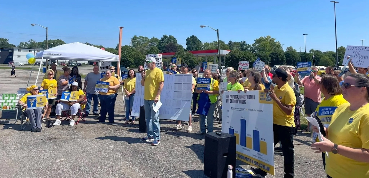 A group in a parking lot in yellow t-shirts listen while one man speaks through a microphone
