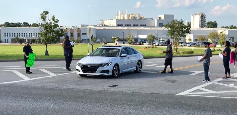 Workers outside a big plant approach a car with leaflets.