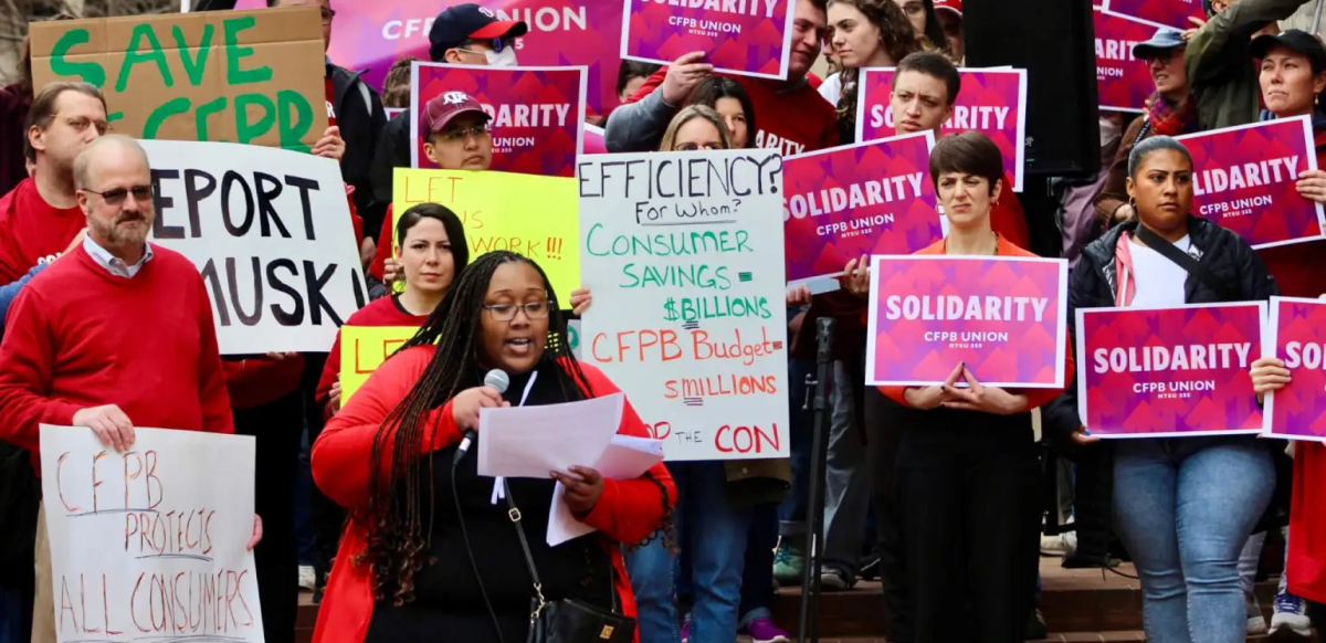CFPB workers at a rally holding pink signs that say Solidarity and other handmade signs with slogans like CFPB Protects All Consumers.