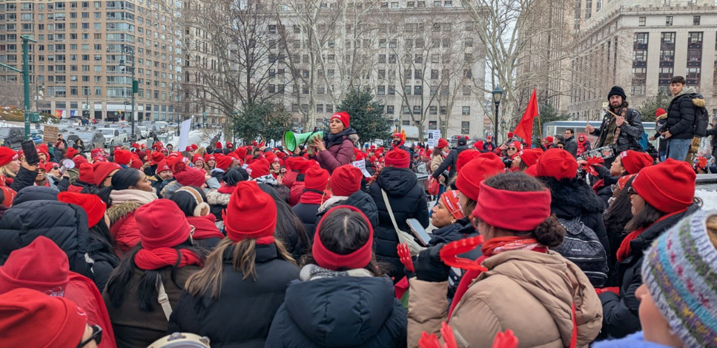 A sea of people, mostly women, of various races, bundled up in winter clothes with red hats and scarves, packs a square in New York City. One person with a megaphone stands elevated a bit in the center, surveying the crowd.