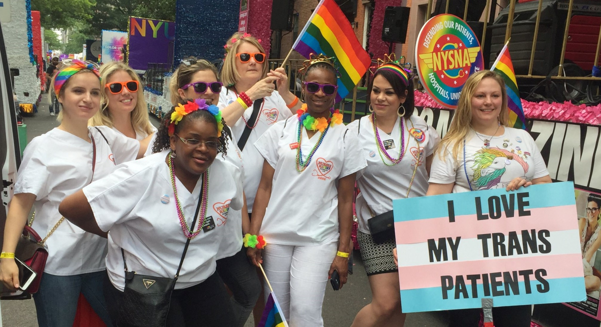 Nurses in white scrubs with rainbow flags and accessories; one carries a sign "I Love My Trans Patients" with the trans flag (pastel blue, pink, and white stripes)