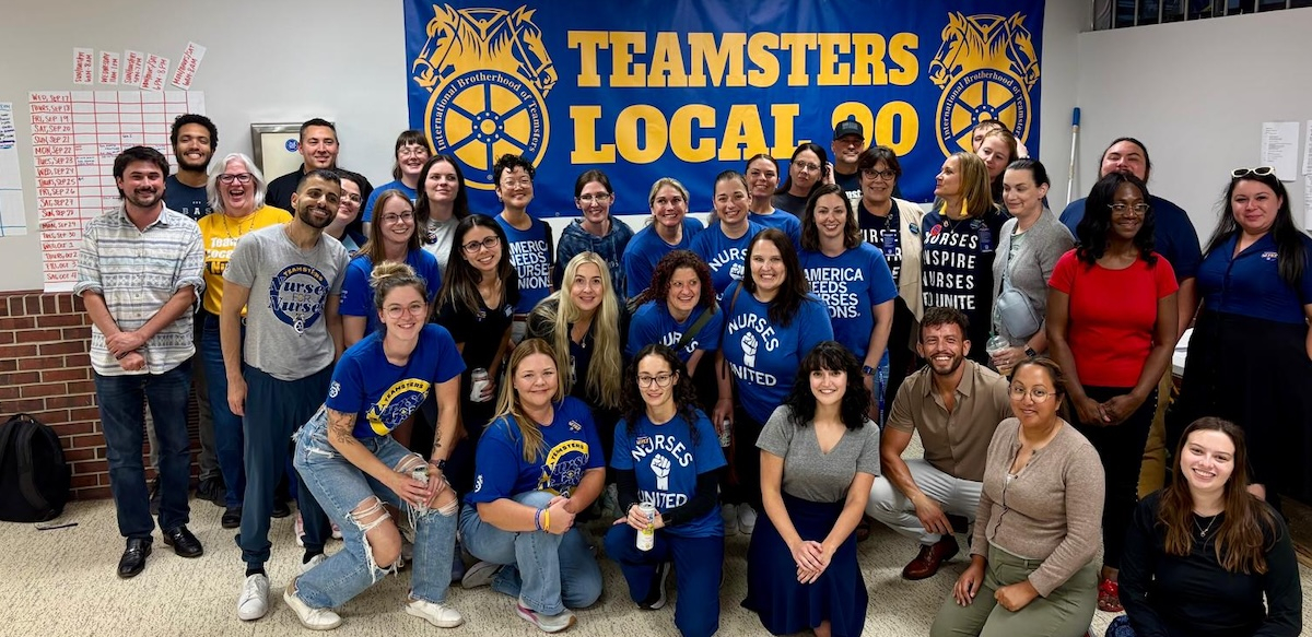 A group of 25 blue-shirted nurses pose happily in front of a Teamsters Local 90 banner
