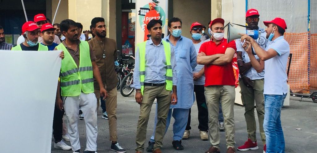 A group of mainly Pakistani immigrant workers stand at a rally while a union organizer speaks through a bullhorn.